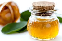 Small glass jar with golden honey and cork stopper on white background with green leaves, close up view showing liquid texture and transparency.