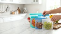 Young adult with medium skin tone organizing food storage containers with fresh produce and cereals in a bright kitchen setting for meal prep and pantry organization.