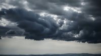 Dramatic dark storm clouds gathering in overcast sky with rays of sunlight breaking through heavy gray cloudscape before thunderstorm weather system.