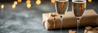 Two elegant champagne flutes filled with sparkling wine stand beside wrapped gift with golden ribbon against warm bokeh lights background for celebration.