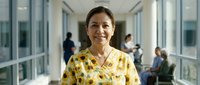 Smiling middle-aged Asian woman in floral blouse standing in modern office corridor with colleagues in background for business and workplace concepts.