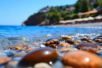 Smooth wet pebbles on Mediterranean beach with crystal clear water, coastal houses and green hills in soft focus background, summer seascape.