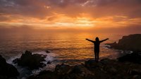 Person with arms outstretched celebrating freedom on rocky coastline during golden sunset over ocean waves with dramatic orange sky clouds.