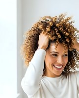Happy mixed race woman with curly hair touching head and smiling in white sweater for beauty and lifestyle content.