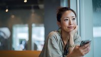 Thoughtful Asian businesswoman holding smartphone in modern office environment, contemplating future plans and digital communication strategies.