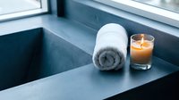 Rolled white towel and lit candle on modern bathroom sink counter near window, spa and wellness relaxation setting.