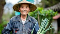 Elderly Asian farmer wearing traditional conical hat holds fresh lemongrass stalks from harvest, smiling warmly in casual work clothes against blurred garden background.