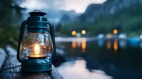 Vintage oil lantern with glowing flame against blurred mountain lake at dusk, warm lights reflecting on water surface creates peaceful outdoor atmosphere.