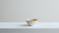 White ceramic bowl with golden liquid on minimalist white surface against neutral gray background, modern clean composition for food and beverage styling.