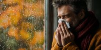Middle-aged Caucasian man with beard blowing nose into tissue by rainy window with autumn colors outside, wearing scarf and sweater during cold season.