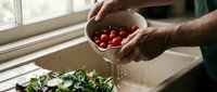 Man with dark skin tone washing fresh red cherry tomatoes in a white colander over a kitchen sink with green salad nearby, food preparation and healthy cooking concept.