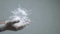 Hand holding glowing coronavirus model with rainbow light effects against gray background for medical education and healthcare concepts.