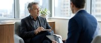 Mature male professional with gray hair in dark blazer takes notes during business meeting with colleague in modern office with city skyline view, corporate consultation.
