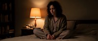 Young woman sitting on bed in cozy bedroom with warm lamp light, wearing comfortable pajamas, evening relaxation and peaceful nighttime atmosphere.