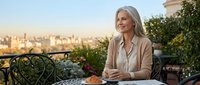 Mature blonde businesswoman enjoying morning coffee on urban terrace overlooking city skyline during golden hour sunrise.