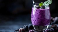 Purple smoothie with frozen blackberries and mint garnish in glass on dark background, fresh berries scattered around for healthy breakfast drink.