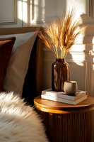 Golden wheat stalks in amber glass bottle on wooden nightstand with books and candle, warm sunlight creating cozy bedroom atmosphere.