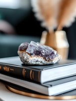Raw amethyst crystal cluster on stack of books with soft focus background, natural purple gemstone specimen displaying crystalline formation and healing properties.