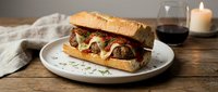 Meatball sandwich with melted cheese on toasted bun, served on white plate with wine and candle for upscale dining or restaurant menu photography.