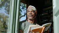 African American man reading book outdoors on porch wearing casual clothing and headband, enjoying leisure time learning and education at home.