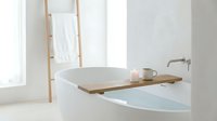 Minimalist bathroom interior with wooden bathtub caddy holding candles and mug, white towel on ladder, bright natural light, spa relaxation setting.