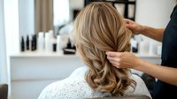 Professional hairstylist creating elegant wavy hairstyle for blonde woman in salon, back view with soft natural lighting and blurred cosmetic products.