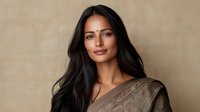 Young South Asian woman with long dark hair wearing traditional brown sari and bindi, smiling confidently against neutral beige background. Professional portrait with soft lighting.