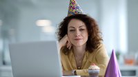 Woman with curly hair wearing colorful party hat smiles at laptop screen with cupcake and purple party hat on desk, celebrating birthday remotely in bright office setting.