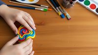 Child hands painting colorful rainbow heart shape with watercolors on wooden table surrounded by art supplies and brushes for creative projects.