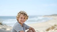 Young boy with curly blonde hair and fair complexion smiling at beach with ocean and sandy shore in background, suitable for family, lifestyle, and travel content.