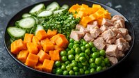 Fresh bowl with diced butternut squash, green peas, cucumber slices, chopped parsley, and cooked chicken pieces arranged on dark plate against stone surface.