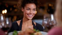 Young African American woman with curly hair smiling warmly in restaurant setting with wine glasses and ambient lighting, expressing genuine happiness and hospitality.