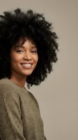 Happy young African American woman with natural curly afro hair smiling warmly in cozy knit sweater against neutral beige background for lifestyle content.