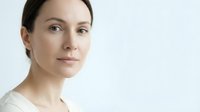 Professional portrait of confident young Caucasian woman with natural makeup and brown hair pulled back, wearing white top against clean background.