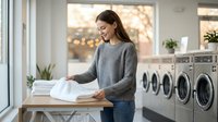 Young woman folding clean white laundry in modern laundromat with washing machines. Domestic chores and household tasks concept for lifestyle content.