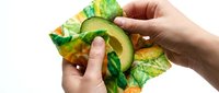 Hands wrapping a fresh avocado half in reusable beeswax food wrap with green leaf pattern, eco-friendly zero waste kitchen concept.