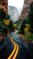 Winding mountain road curves through towering red rock canyon walls with yellow autumn trees and evergreen forest creating dramatic natural scenery.