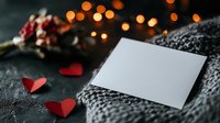 Blank white card mockup with red paper hearts on cozy knitted sweater background with bokeh lights and winter berries, romantic holiday setting.