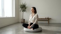 Young Asian woman practicing mindfulness meditation sitting cross-legged on cushion in minimalist room with natural light and plant for wellness.