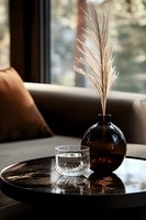 Minimalist home decor with dried pampas grass in dark glass vase and crystal glass on black marble tray, cozy interior setting near window with natural light.