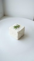 Fresh white cheese block topped with rosemary sprig on clean white surface. Creamy feta or ricotta cube for culinary, food styling, and recipe content.