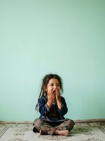 Young child, approximately 3-5 years old, with dark hair and medium skin tone, sitting cross-legged on weathered wooden floor against soft mint green wall, hand covering mouth in surprised or thoughtful expression, wearing dark patterned clothing, suitable for parenting, child development, and emotional expression concepts.