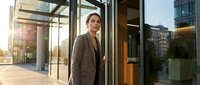 Professional businesswoman in elegant beige coat entering modern glass office building during golden hour sunset, corporate lifestyle and success concept.