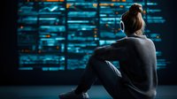 Young Asian female programmer in casual wear with headphones sitting on floor looking at glowing blue code on large screen in dark room. Cybersecurity concept.