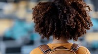 Young African American woman with natural curly afro hair shown from behind wearing yellow sweater and backpack against blurred urban background. Focus on textured hair.