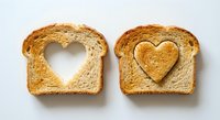 Two slices of golden whole grain bread on white background, one with heart-shaped cutout, other with heart piece placed inside for romantic breakfast concept.