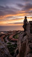 Woman in casual athletic wear standing on rocky cliff edge overlooking coastal town and sea at golden hour sunset, contemplating landscape view for travel, adventure, and lifestyle concepts.