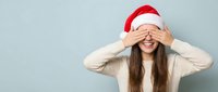 Young woman in Santa hat covering eyes with hands in playful peek-a-boo gesture against light blue background for holiday marketing campaigns.