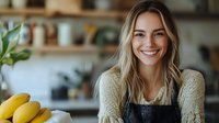 Young Caucasian woman with blonde hair wearing apron and sweater smiling in kitchen with fresh bananas and plants, representing healthy lifestyle and home cooking.