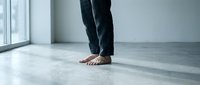 Person with bare feet wearing dark pants standing on polished concrete floor in minimalist interior space with natural light from window.